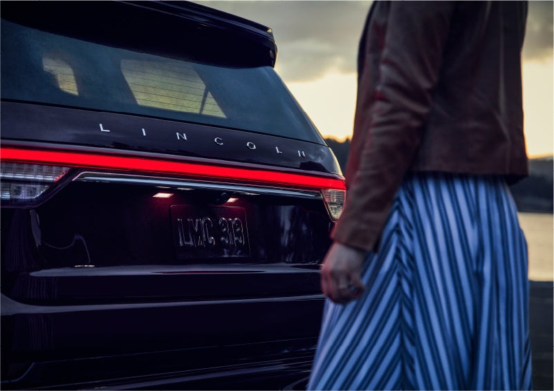 A person is shown near the rear of a 2023 Lincoln Aviator® SUV as the Lincoln Embrace illuminates the rear lights | Hines Park Lincoln in Plymouth MI