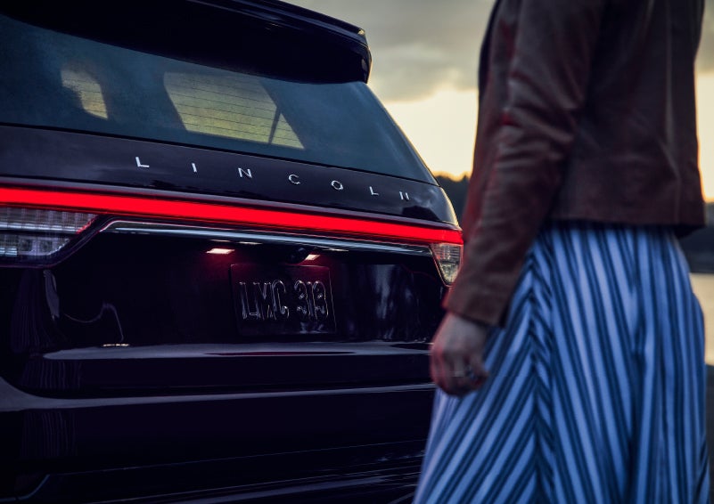 A person is shown near the rear of a 2024 Lincoln Aviator® SUV as the Lincoln Embrace illuminates the rear lights | Hines Park Lincoln in Plymouth MI