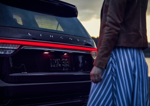 A person is shown near the rear of a 2024 Lincoln Aviator® SUV as the Lincoln Embrace illuminates the rear lights | Hines Park Lincoln in Plymouth MI